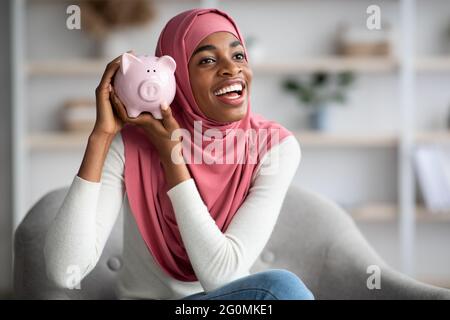 Fröhliche Schwarze Muslimische Frau In Hijab Hält Sparschwein, Während Sie Zu Hause Sitzt Stockfoto