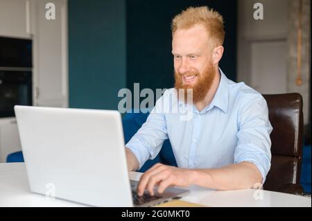 Fröhlicher, rothaariger Hipster-Mann, der für Fernarbeiten einen Laptop benutzt, ein glücklicher junger bärtiger Mann im formellen Hemd, der auf der Tastatur am Schreibtisch im Loft-Heimbüro sitzt Stockfoto