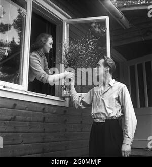 Lifestyle in den 1950er Jahren. Ein junges Paar, das in ihrem Haus abgebildet ist. Durch das offene Fenster reicht er ihr einen Strauß Äste mit kleinen Blättern. Schweden 1951 Ref. BB80-1 Stockfoto