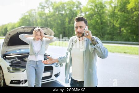 Junger Mann, der in der Nähe eines kaputten Autos mit einer verängstigten Freundin stand, einen Autounfall hatte und den Pannendienst anrief Stockfoto