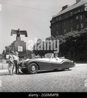 Fahren in den 1950er Jahren. Eine junge Frau in einem offenen Cabriolet des Jaguar XK120. Ein britischer Sportwagen, der als futuristisch anmutet. Die Zahlen 120 geben die Höchstgeschwindigkeit des Fahrzeugs an, 120 mph/193 km/h. Das Auto war ein Zweisitzer mit einer Aluminium-Chasse. Es wurde in insgesamt 12078 Autos hergestellt. Auf der Straße vom Flugdenkmal von Carl Milles auf Karlaplan in Stockholm abgebildet. Eine Hommage an schwedische Piloten und geflügelte Pioniere. Schweden 1953. ref. BL108-12 Stockfoto