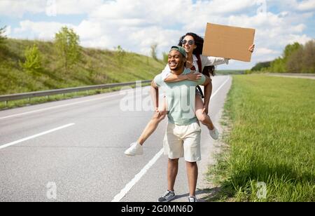 Tausendjähriges multirassisches Paar, das mit einem Autostop unterwegs ist, mit Anhalter unterwegs ist, nach Transport sucht und ein leeres Schild mit Mockup hält Stockfoto