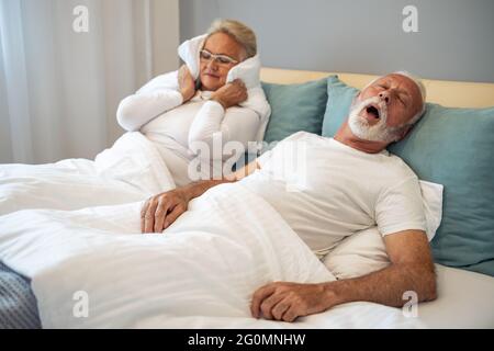 Gestresste ältere Frau, die ihre Ohren bedeckt, weil ihr Mann schnarcht.Ältere Paare haben Probleme mit dem Schnarchen des Mannes. Stockfoto