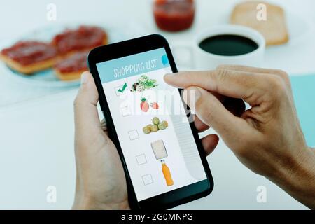 Nahaufnahme eines jungen kaukasischen Mannes, der zum Frühstück an einem Tisch sitzt, seine Einkaufsliste auf seinem Smartphone mit Bildern verschiedener Produkte erstellt Stockfoto