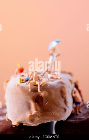 Eine Gruppe von Miniatur-Männern und -Frauen, die Badeanzug tragen, und eine alte Dame, die einen Regenschirm trägt, sich an einigen Schokoladen-Eisdielen entspannt, gegen ein Braun Stockfoto