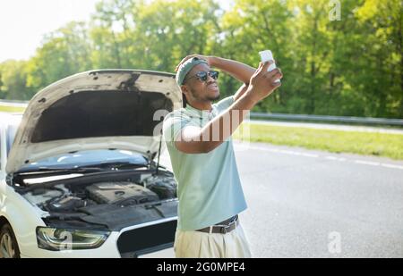 Verärgert schwarzen Kerl in der Nähe gebrochenen Auto mit offener Haube, Probleme, versucht, mobiles Netzwerk zu finden, ohne Signal Stockfoto