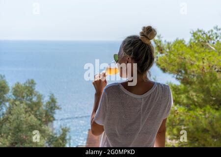 Hübsche Frau im weißen Hemd trinkt Aperol Spritz und genießt den Blick auf das offene Meer Stockfoto