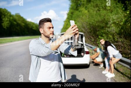 Junger Mann, der auf der Straße neben einem kaputten Auto steht, auf der Suche nach einem Mobilfunksignal ist und versucht, den Pannendienst anzurufen Stockfoto