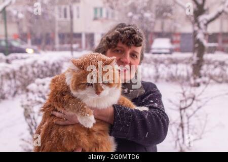 Frau mit lockigen Haaren, die an einem verschneiten Tag eine orangefarbene Katze hält Stockfoto