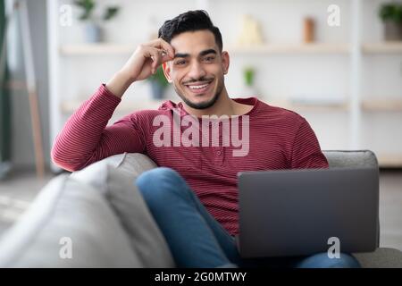 Positiver arabischer Kerl, der sich auf der Couch mit Laptop zurücklehnen kann Stockfoto