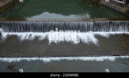 Ein breiter, aber flacher Fluss mit einer Betonwand auf beiden Seiten und einer Betonwand, die in der Mitte einen kleinen Wasserfall bildet. Stockfoto