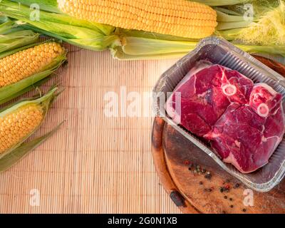 Flache Lage von gelben Maiskolben mit grünen Blättern auf einem Tisch und in großen Topf und rohem Rindersteak in einer Folienpfanne und ganzen Pfeffer auf einem hölzernen Schneidebrett. Stockfoto