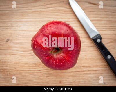 Frischer, saftiger, nasser roter Apfel mit Wassertropfen auf der Haut und einem Messer auf einem Holzschneidebrett. Apple a day gesundes Konzept. Stockfoto