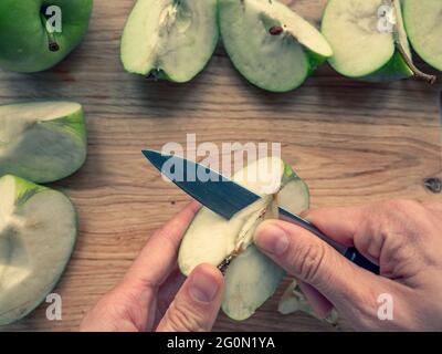 Hand mit Messer, die einen Apfelkern auf einem hölzernen Schneidebrett ausschneiden, frische saftige grüne Apfelviertel auf dem Hintergrund verstreut. Stockfoto