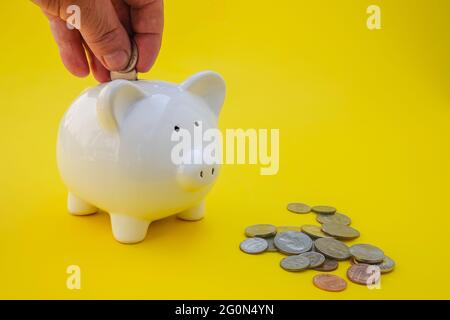 Mann, der Münzen in ein weißes Sparschwein mit amerikanischen Münzen auf gelbem Hintergrund legt Stockfoto