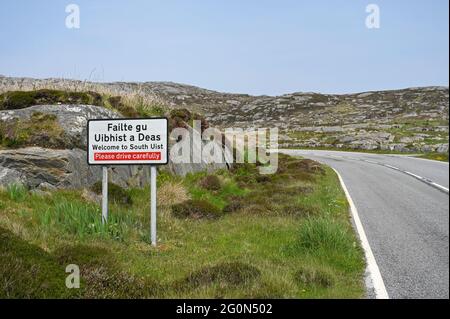 Willkommen beim South Uist-Zeichen auf Englisch und Schottisch-Gälisch. Hintergrund von Straße, Gras und felsiger Landschaft. Sonniger Tag mit blauem Himmel, keine Menschen. Stockfoto