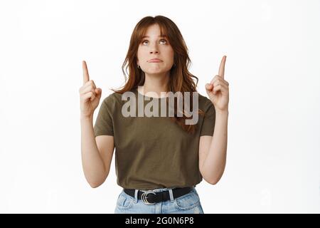 Trauriges und düsteres Mädchen zeigt die Finger nach oben, schaut auf Top-Werbung mit pouting verärgert Gesicht, zum Ausdruck bringen Bedauern, wollen etwas zu bekommen, stehen gegen Stockfoto