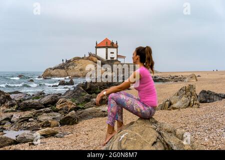 Junge Frau, die auf einem Felsen am Strand sitzt und in die Ferne blickt. Kapelle Senhor da Pedra am Strand Miramar, Vila Nova de Gaia, Porto im Hintergrund Stockfoto