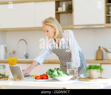 Fokussierte ältere Frau in Schürze, die im Internet Video Rezept auf digitalem Tablet beim Kochen in der Küche zu Hause, frisches Gemüse auf dem Tisch. Aktive pensionierte Frau, die das Leben nach der Pensionierung genießt Stockfoto