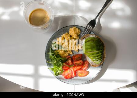 Teller mit Rührei, Rucola, Tomate, Vollkorntoast mit Avocado, Tasse Kaffee auf dem Tisch. Stockfoto