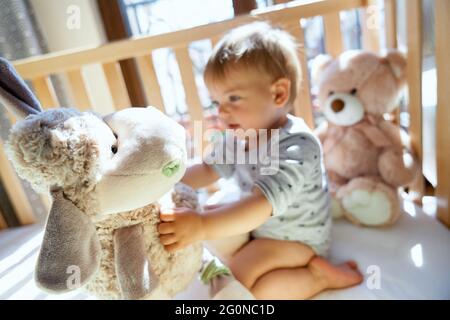 Lächelndes kleines Kind, das auf einem Holzbett sitzt und ein großes Plüschschaf in der Hand hält Stockfoto