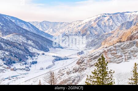 Winter verschneite Landschaft des Tales zwischen den hohen felsigen Bergen mit einer Autobahn, Straße und einer Dorfsiedlung mit einer Brücke über den Fluss Stockfoto