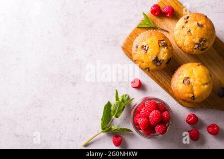 Frisch gebackene Cupcakes auf Holzbrett neben Himbeeren und frische Minze auf grauer Arbeitsplatte Stockfoto