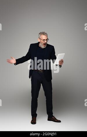 Ganze Länge der wütend mittleren Alters Geschäftsmann mit grauen Haaren schreiend, während Blick auf digitale Tablet auf grau Stockfoto
