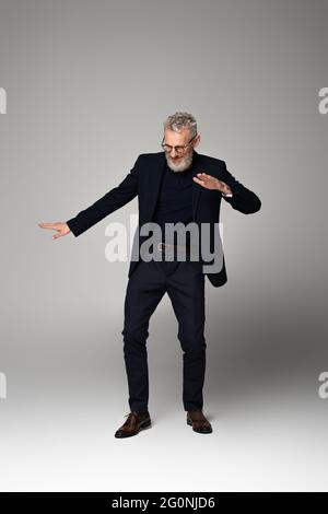 Die ganze Länge des fröhlichen Mannes mittleren Alters in Gläsern und Anzug tanzen auf grau Stockfoto