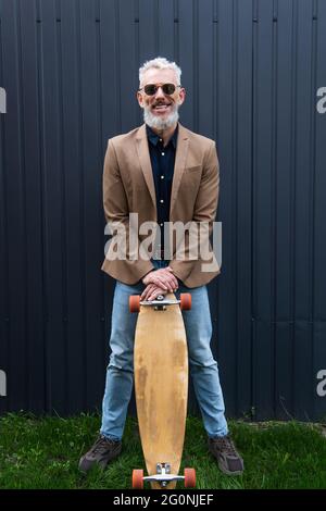 Die ganze Länge eines glücklichen Mannes mittleren Alters in einer Sonnenbrille, der Longboard draußen hält Stockfoto