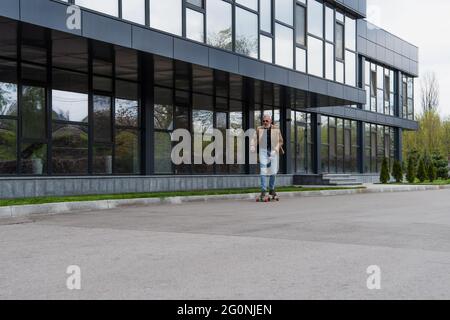 Die ganze Länge des bärtigen und reifen Mannes in Sonnenbrillen Reiten Longboard draußen Stockfoto