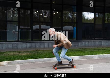 Die ganze Länge des bärtigen Mannes mittleren Alters in Sonnenbrillen, der draußen auf dem Longboard reitet Stockfoto