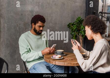 Junges afroamerikanisches Paar mit Smartphones in der Nähe von Makronen und Kaffee im Café Stockfoto