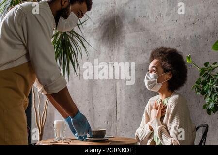 afroamerikanischer Klient in medizinischer Maske sitzt in der Nähe von Barista mit Kaffee im Café Stockfoto