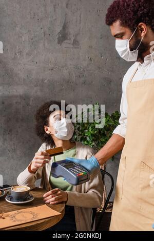 afroamerikanischer Kunde mit Kreditkarte in der Nähe des Barista in einer medizinischen Maske mit Zahlungsterminal Stockfoto