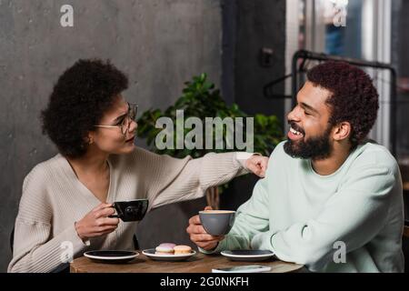 Seitenansicht eines lächelnden afroamerikanischen Mannes mit Kaffee, der in der Nähe einer wütenden Freundin im Café sitzt Stockfoto