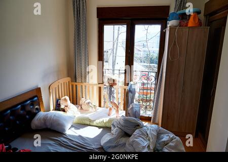 Das kleine Kind steht in einem Kinderbett neben einem Erwachsenenbett vor einer Balkontür Stockfoto