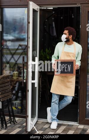 afroamerikanischer Barista in medizinischer Maske mit einer Tafel mit offenen Schriftzügen im Café Stockfoto