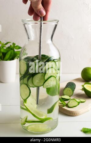 Eine Hand rührt mit einem Metallstroh die Zutaten in einem Krug mit frischem Wasser, das mit Gurken und Minzspalten, einem Detox-Getränk, angereichert ist. Vertikale oris Stockfoto