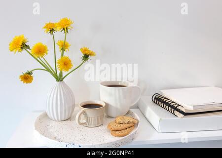 Ein Bouquet von Delelionen in einer weißen, geriffelten Vase und zwei Tassen in verschiedenen Größen mit Kaffee auf einem weißen Tisch. Frühstück. Speicherplatz kopieren Stockfoto
