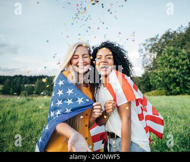 Zwei glückliche Frauen unter der Flagge der USA. Junge Freunde feiern den 4. Juli und werfen Konfetti auf ein Feld. Stockfoto
