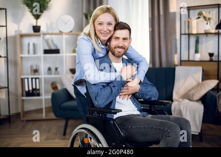 Porträt eines glücklichen Ehepaares, das lächelt und die Kamera im Wohnzimmer anschaut. Hübsche Frau umarmt ihren behinderten Mann. Positive Stimmung. Stockfoto