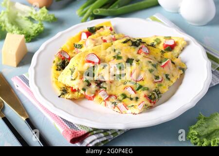 Omelette mit Krabbenstäbchen, Käse und grünen Zwiebeln auf einem weißen Teller auf hellblauem Hintergrund. Nahaufnahme. Stockfoto