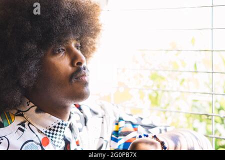 Nahaufnahme eines jungen schwarzen Mannes mit Afro-Haaren und Schnurrbart im Profil neben einem Fenster mit defokussiertem Metallgrill Stockfoto