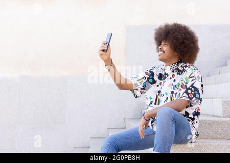 Schwarzer Kerl mit afro Haaren lächelnd sitzt auf einigen Steinstufen und nimmt ein Selfie mit seinem Smartphone Stockfoto