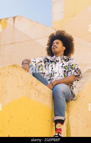 Vertikales Bild von schwarzem Kerl mit afro Haar sitzt auf einem gelben Beton Bordstein mit aufsteigenden diagonalen Linien Stockfoto