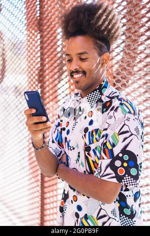 Vertikales Bild von schwarzem Kerl mit afro Haar zurückgezogen Blick auf sein Smartphone auf sonnenbeleuchteten roten Metall Gitter Hintergrund Stockfoto