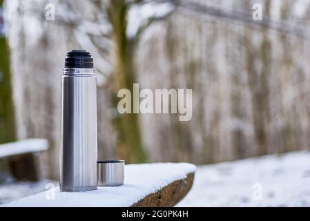 Eine Edelstahl-Thermoskanne mit heißem Tee steht auf einer Bank im Schnee. Winter unfocused Wald Hintergrund. Stockfoto