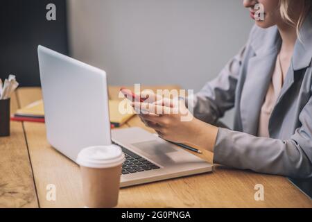 Weibliche Hände halten das Smartphone in der Nähe eines offenen Laptops Stockfoto