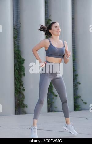 Erhabene Frau, die auf einem Betonpflaster trainiert und mit gespreizten Beinen springt. Über Stützen. In grauem Oberteil und Yogahose. Stockfoto